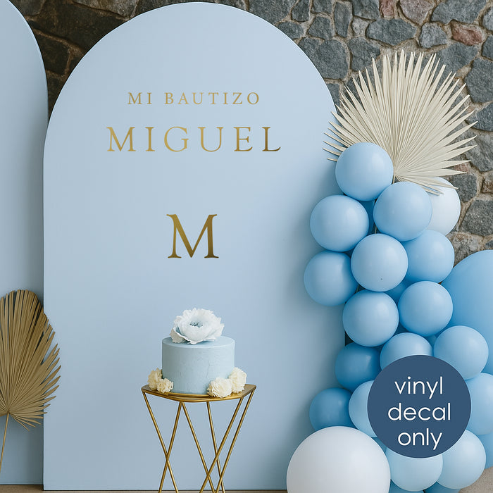 Minimalist gold vinyl decal “Mi Bautizo Miguel” with initial “M” on light blue backdrop for baptism celebration decoration